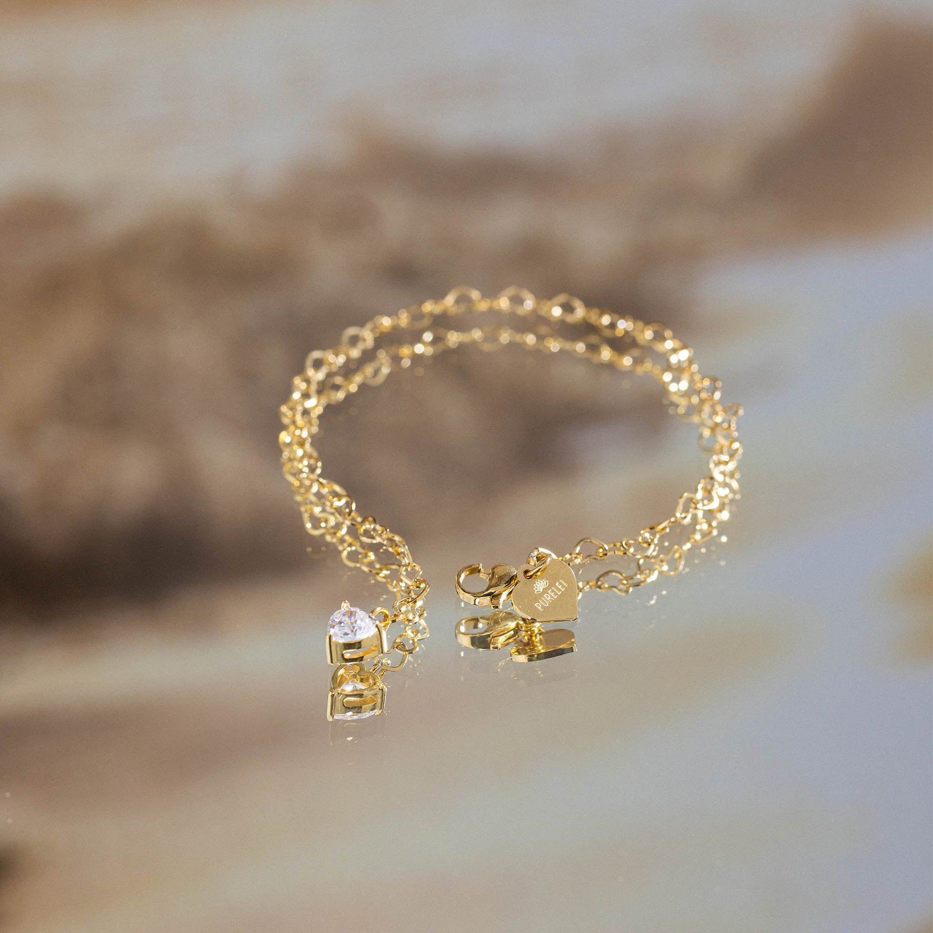 Endloses PURELEI Endless Love Armband in 18K Gold mit Zirkonia-Herzanhänger, elegant auf spiegelnder Oberfläche.