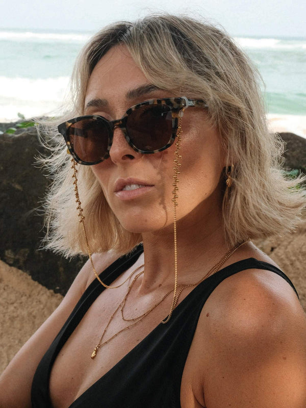 Moderne, braune Sonnenbrille mit Kette, getragen von einer Frau am Strand – PURELEI Stil.