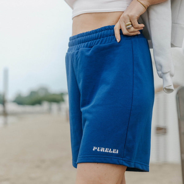 Blue Paradise Shorts aus dunkelblauer Baumwolle mit PURELEI-Logo, sommerlich und bequem getragen am Strand.