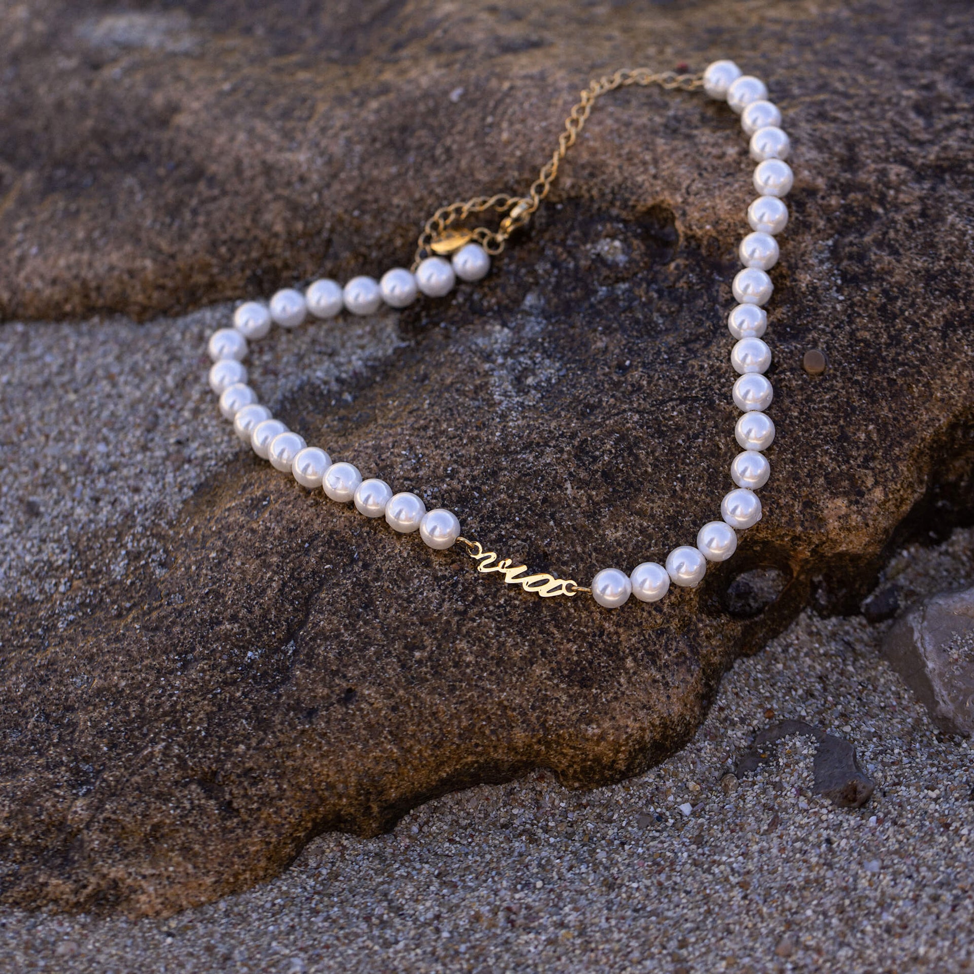 Via Pearl Perlenkette von PURELEI mit goldfarbenem via-Schriftzug und weißen Muschelperlen auf sandigem Stein.