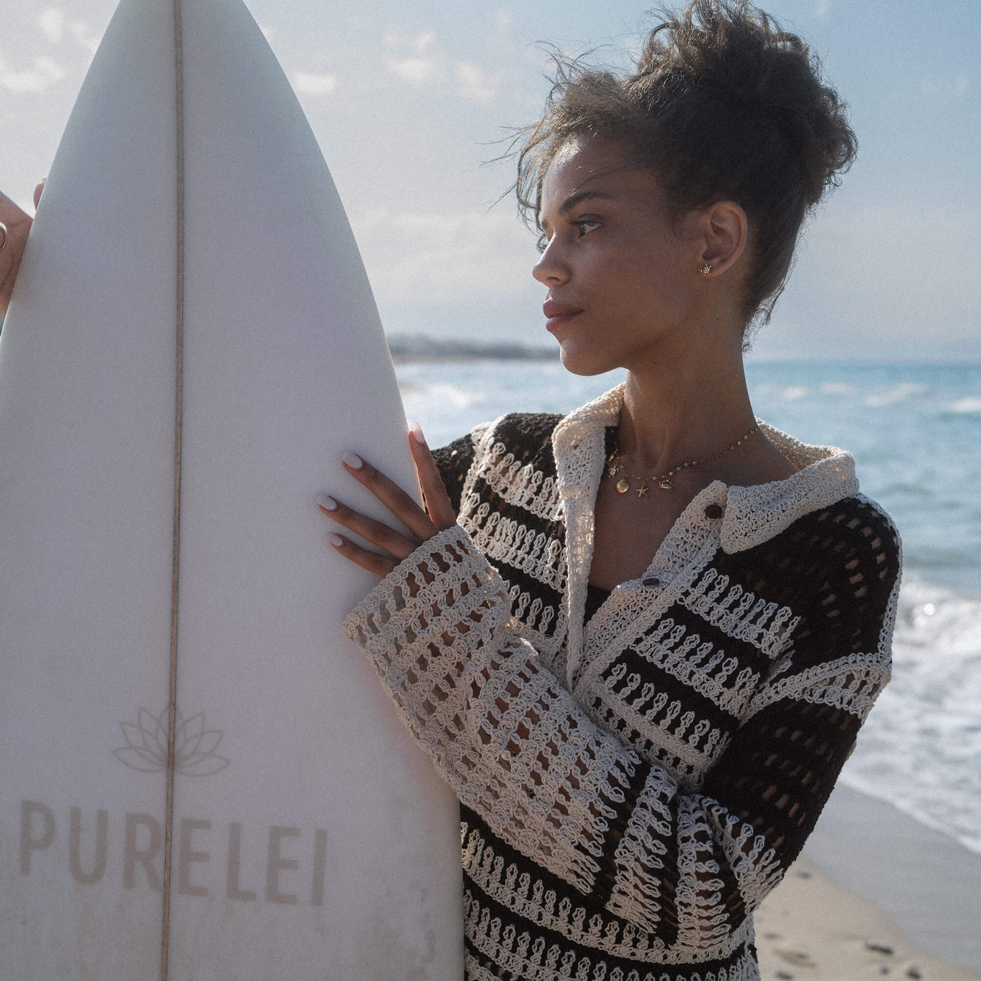 Frau mit schwarzer, gestreifter Strickjacke am Surfbrett, PURELEI-Logo sichtbar.