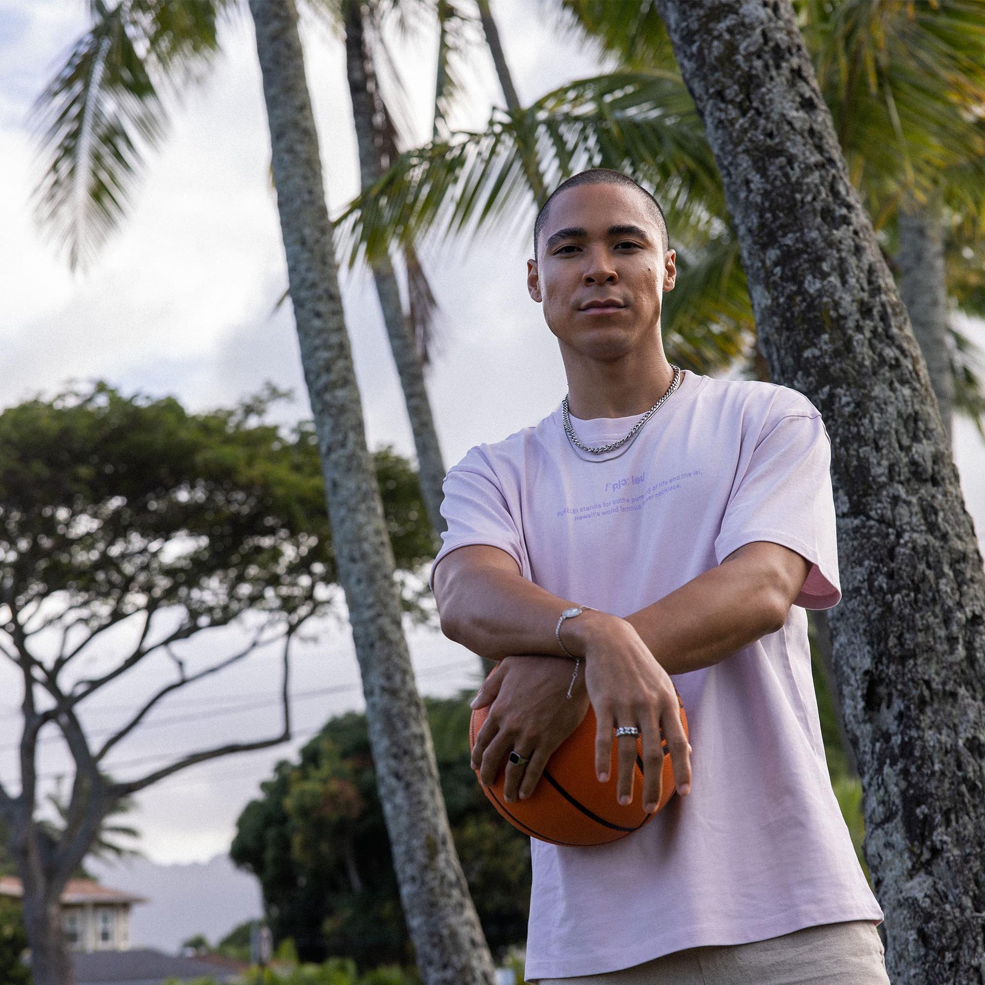 Mann mit Basketball, trägt PURELEI Schmuck, umgeben von Palmen, entspannte tropische Stimmung.