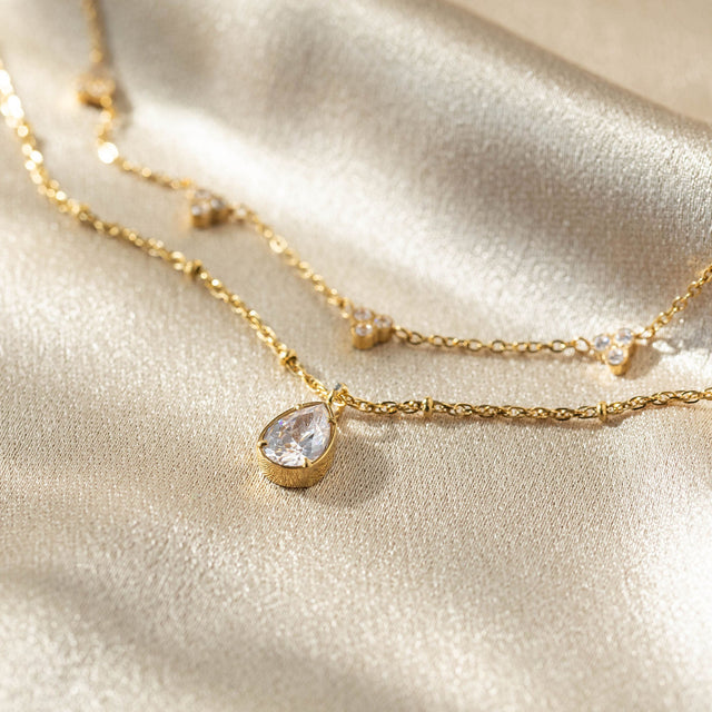 Felice Kette von PURELEI, goldene Edelstahlkette mit weißem Zirkonia-Tropfen, elegant auf schimmerndem Stoff gelegen