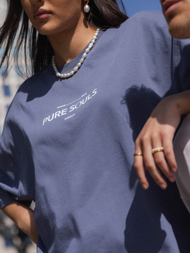 Lila T-Shirt mit PURELEI Schriftzug, kombiniert mit Perlenkette und Ringen, lässige Eleganz.