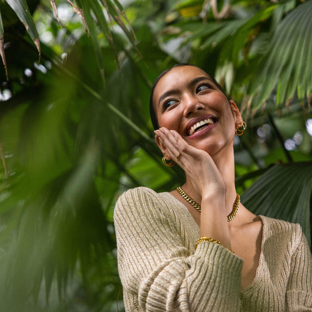 Elegante, goldene Kette und Ohrringe von PURELEI, umgeben von tropischem Grün.