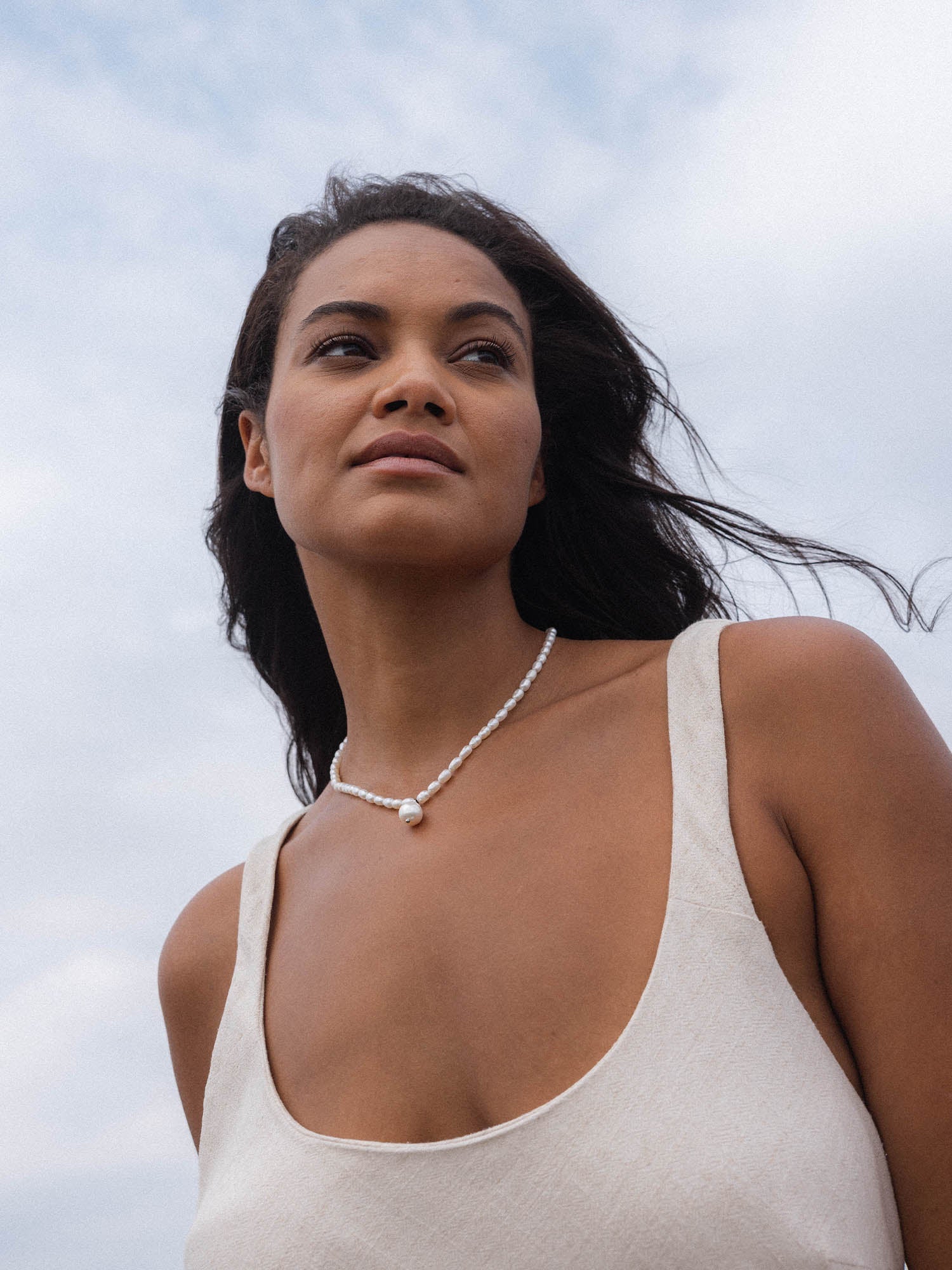 Perlenkette von PURELEI, elegant und feminin, getragen am Strand unter wolkigem Himmel.