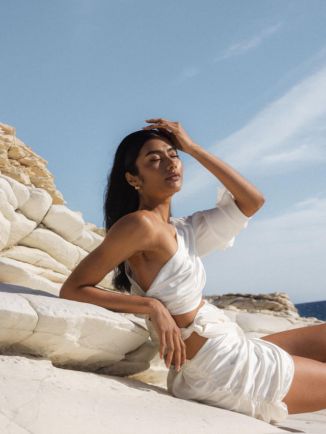 Model mit PURELEI Pisces Letter Charm in Gold, sonnendurchflutet und elegant am Felsen mit Meereshintergrund.