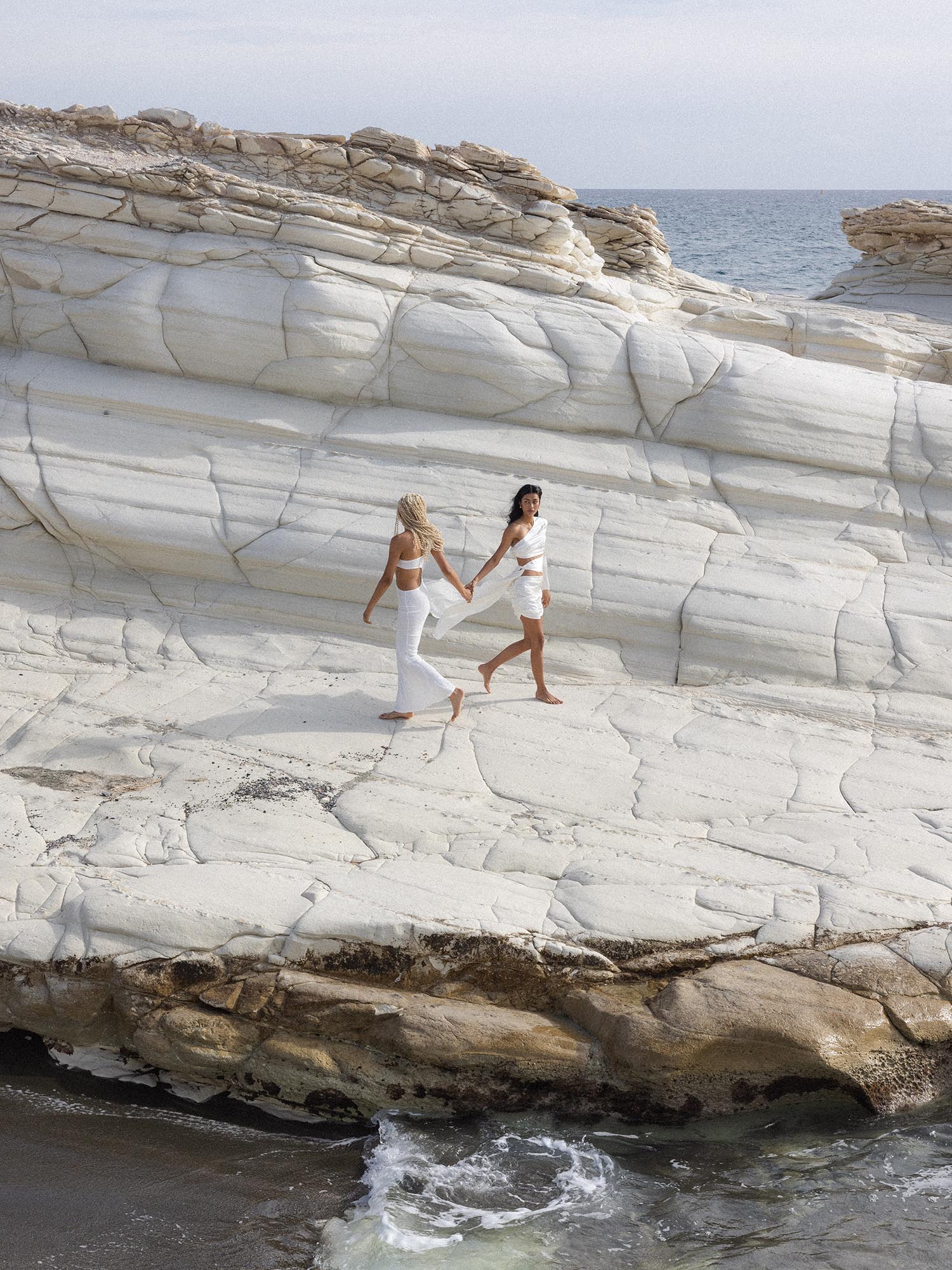 Zwei Frauen laufen barfuß am Strand, beide tragen PURELEI Schmuck, leichte Sommerkleidung, natürliche Eleganz, Meereshintergrund
