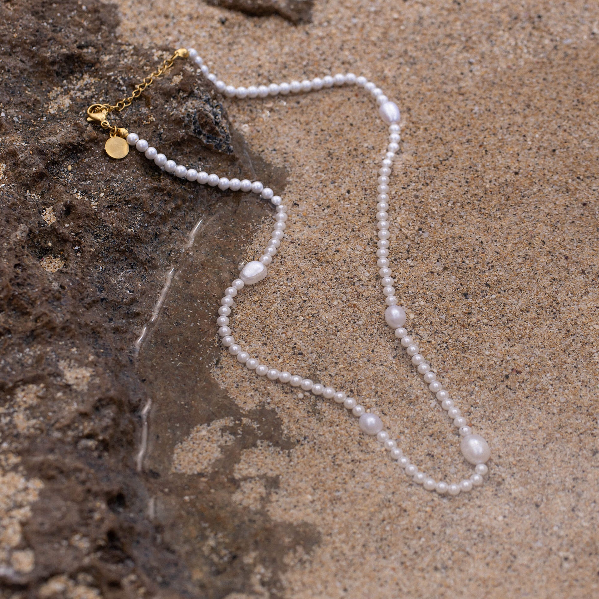 Glace Kette von PURELEI mit Süßwasser- und Muschelperlen, goldfarben, liegt sanft im warmen Sand – elegante Natürlichkeit.