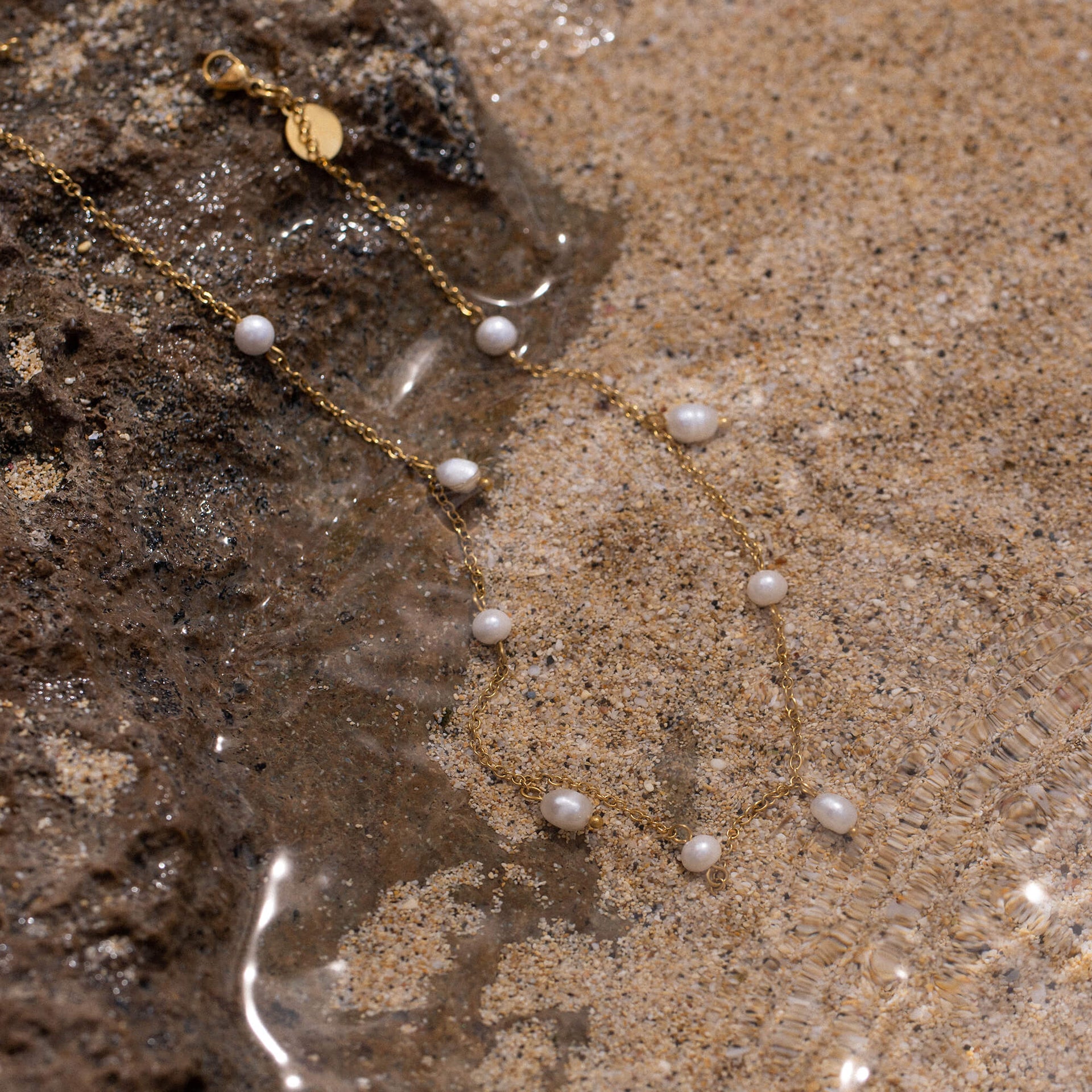 Pearl Drop Fußkettchen von PURELEI mit goldener Kette und zarten Süßwasserperlen, elegant im Sand arrangiert.
