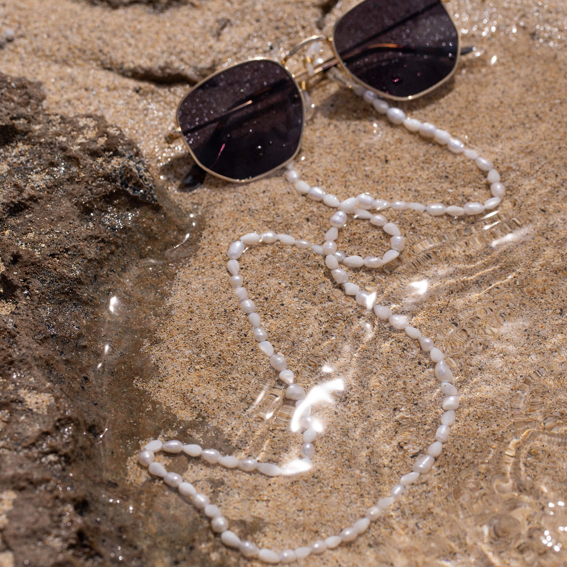 Weiße Muschel-Kette von PURELEI auf Sand, umgeben von Wasser und Sonnenlicht.