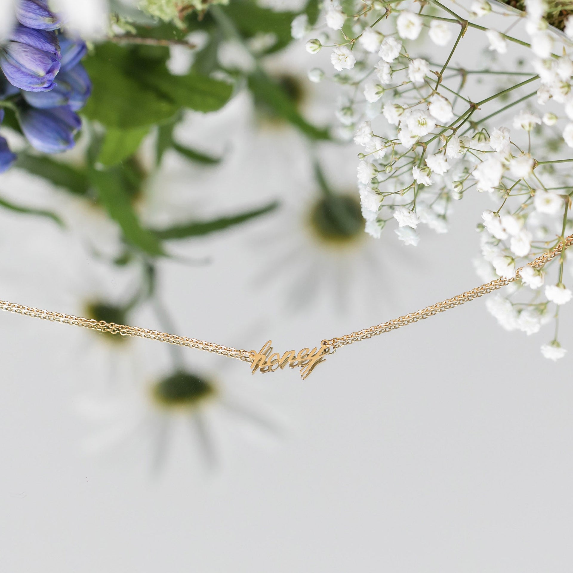 Honey Kette von PURELEI in 18K Gold mit filigranem Schriftzug, elegant inszeniert mit zarten Blüten.